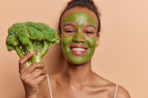 woman smiling with a vegan green mask on holding a broccoli