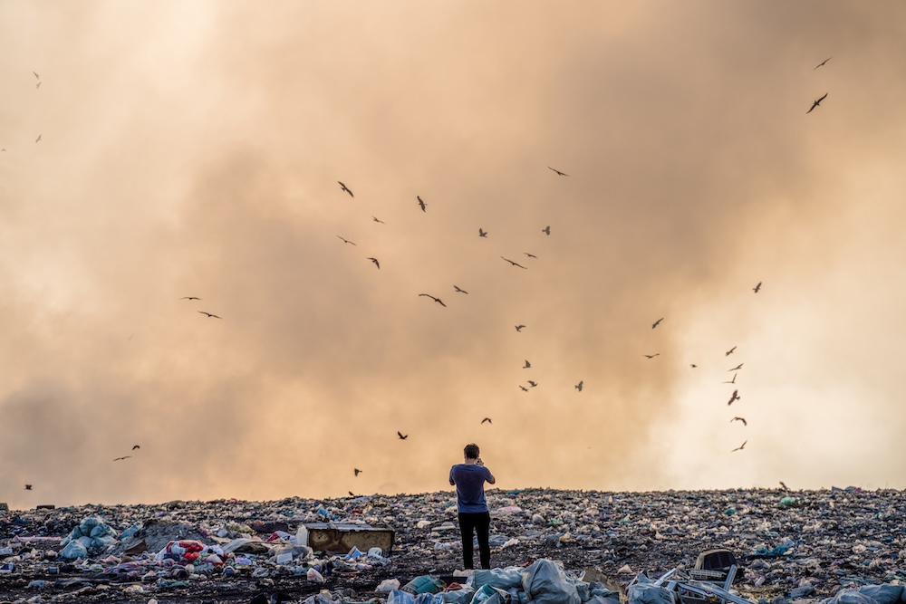 Person standing in the middle of a landfill with hazy sky and birds flying