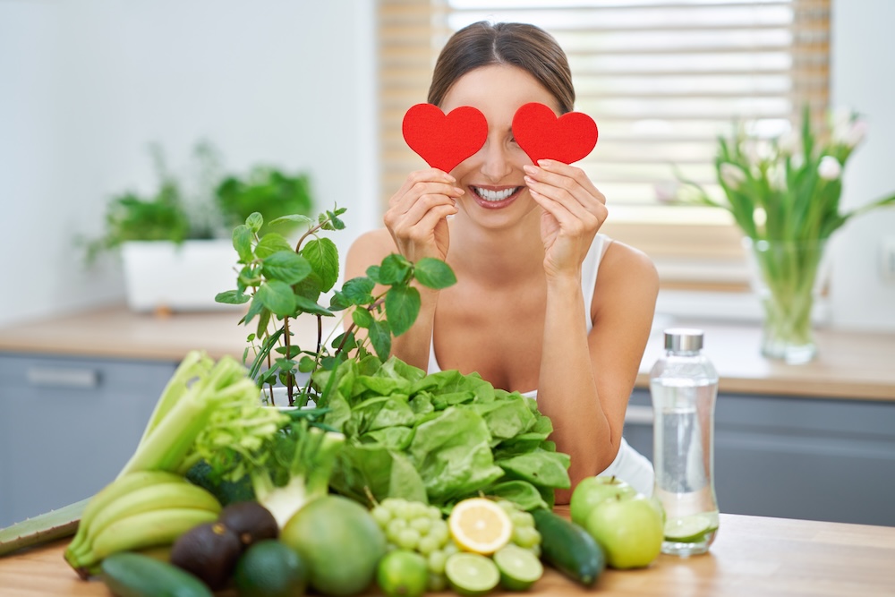 Woman holding cut out red hearts over eyes with leafy greens on table