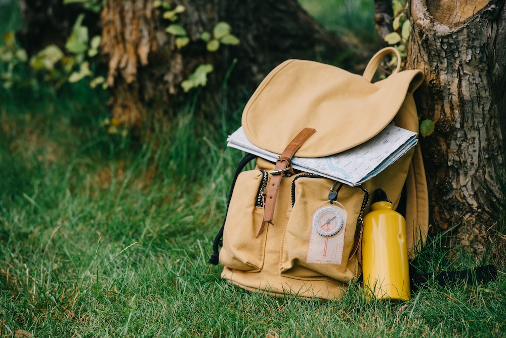 Beige backpack sitting against a tree on the ground in the grass