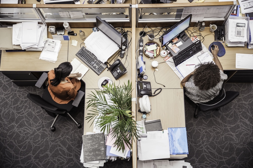 two people sitting at their desks with clutter and waste around them